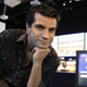 Tommy Tallarico sitting on a table with his hand under his chin, smiling.